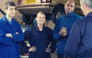 automotive technicians in a shop gathered around being instructed by a teacher